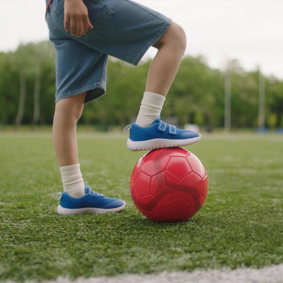 Un copil cu pantaloni scurți și adidași albaștri, cu un picior pe o minge roșie de fotbal.