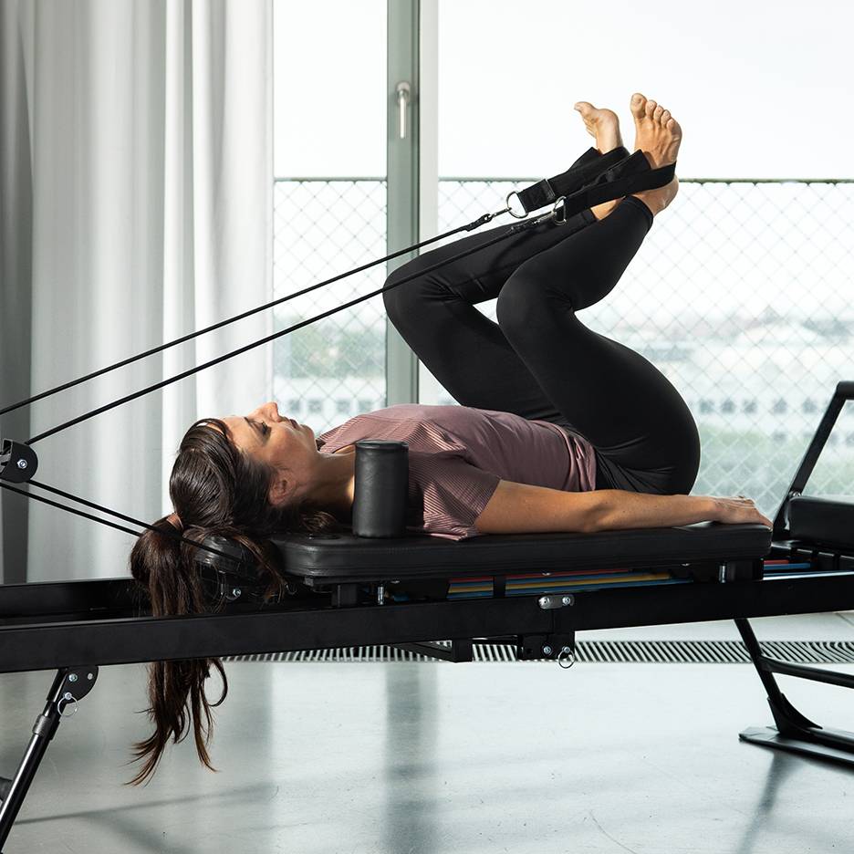 Femeie care își lucrează picioarele pe un aparat Pilates reformer cu benzi de rezistență.