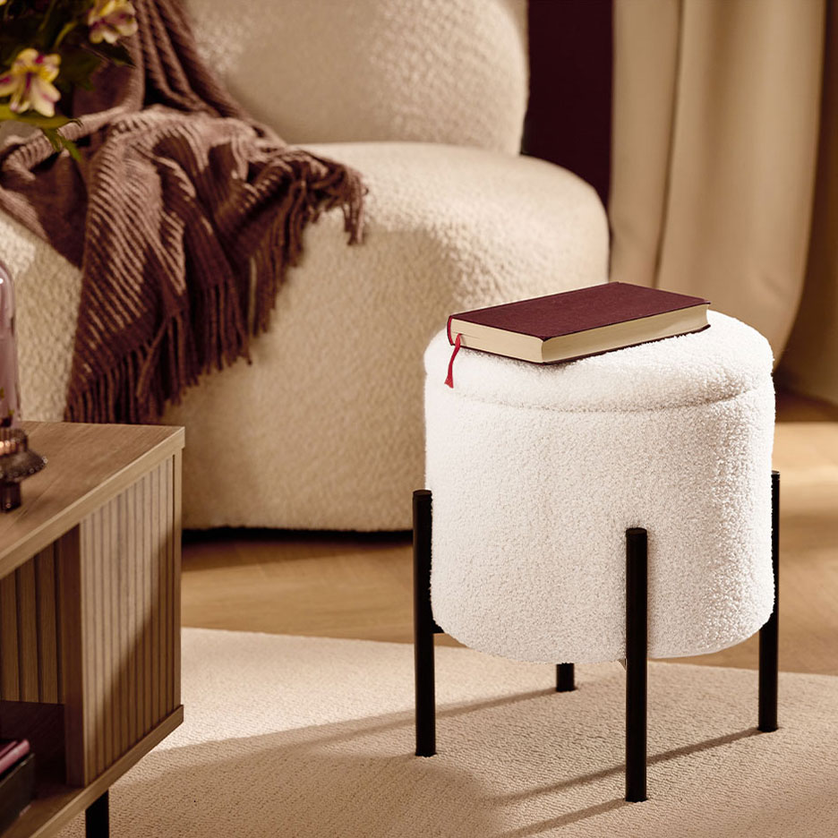 Taburet alb pufos cu picioare negre, o carte roșie, fotoliu și pătură maro.