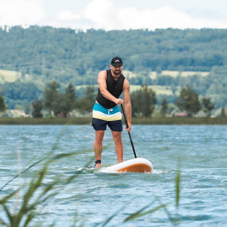Bărbat care face stand-up paddle pe un lac, cu vestă de salvare și pantaloni scurți de baie.