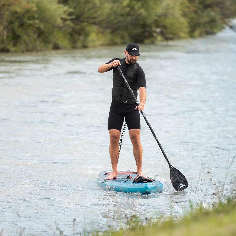 Bărbat face paddleboarding pe un râu, purtând un costum de neopren negru, vestă de salvare și șapcă.