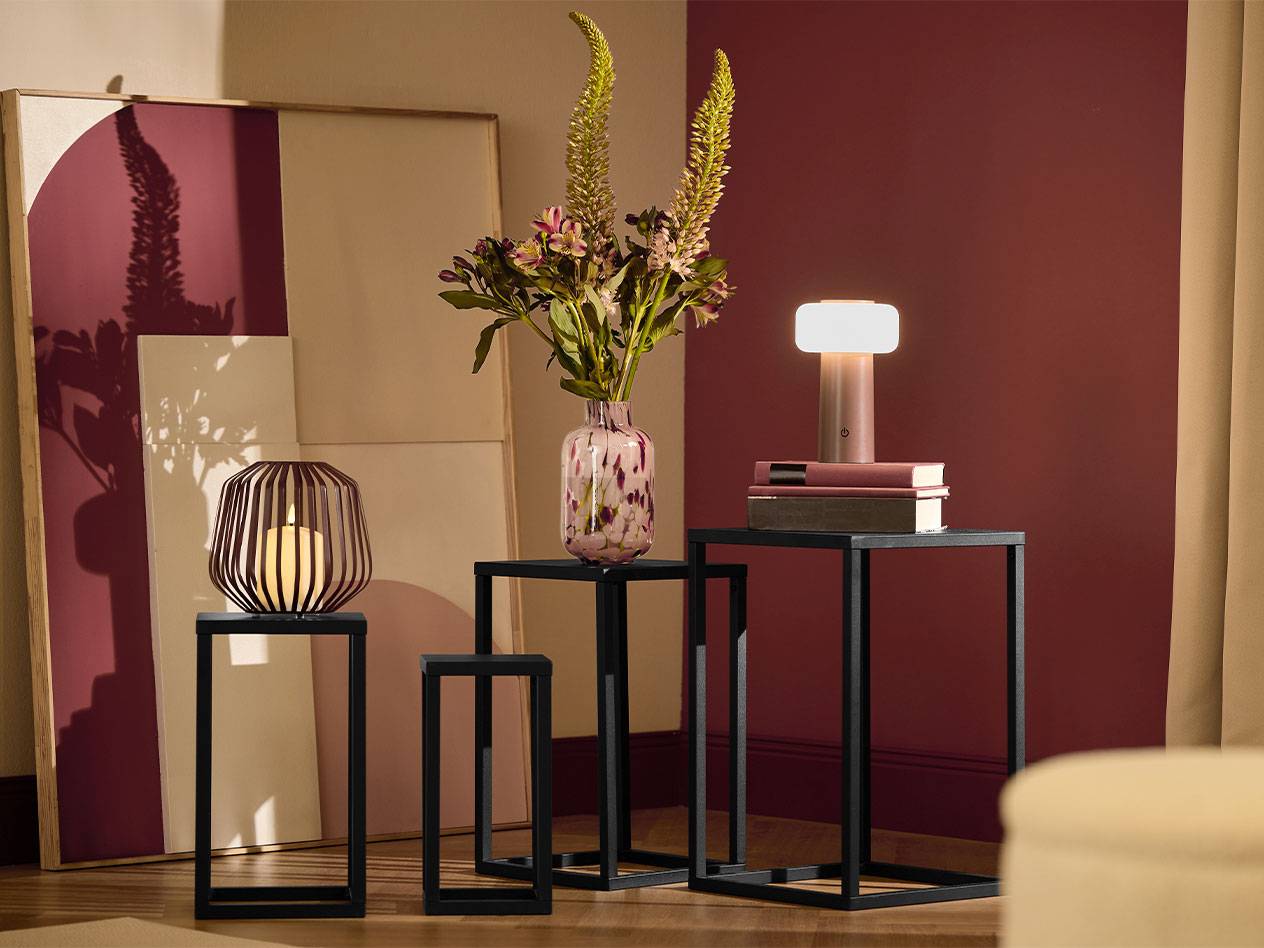 Trei mese laterale negre cu o lumânare, o vază cu flori și o lampă.