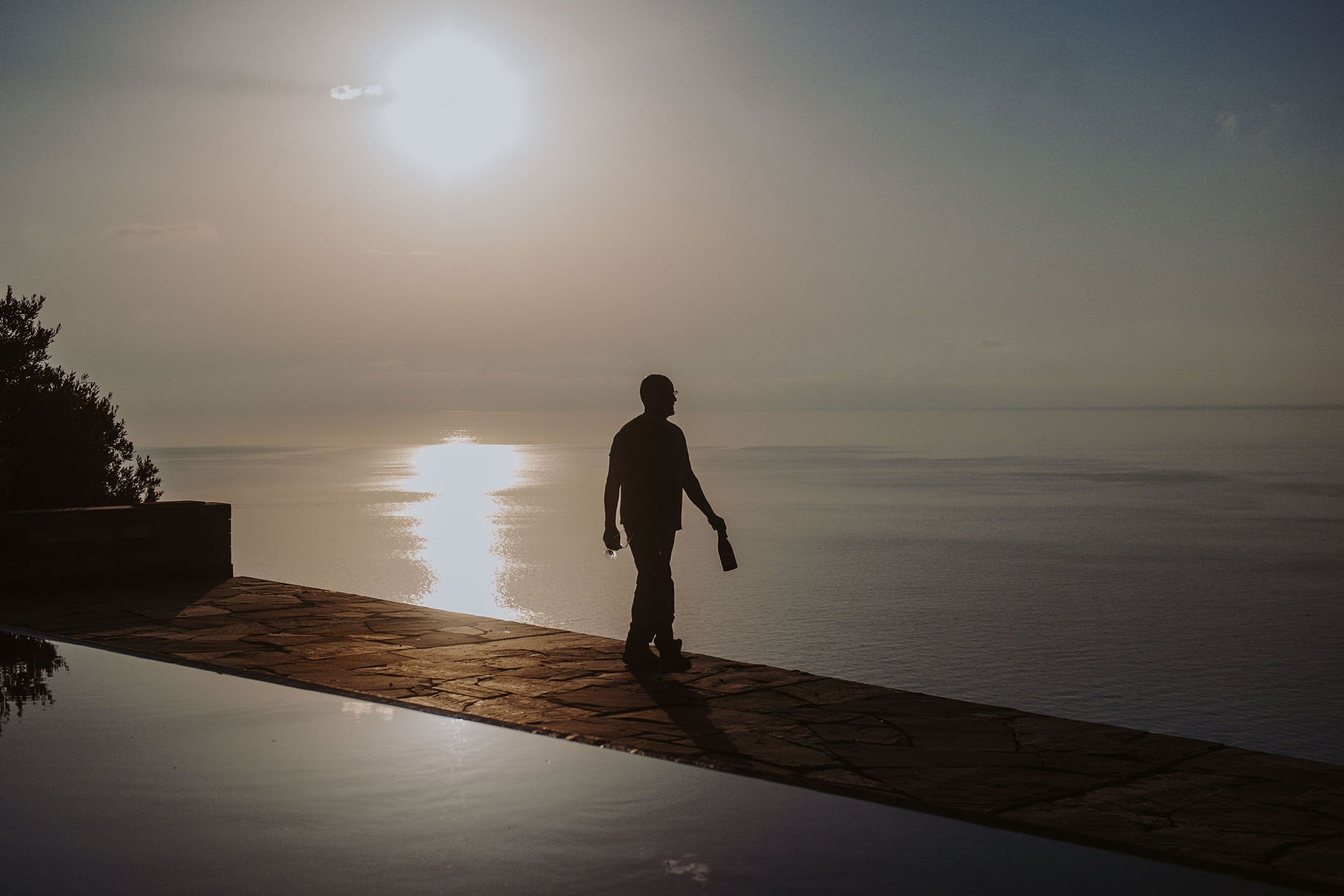 Silueta unei persoane cu sticlă și pahar lângă o piscină infinită la apus