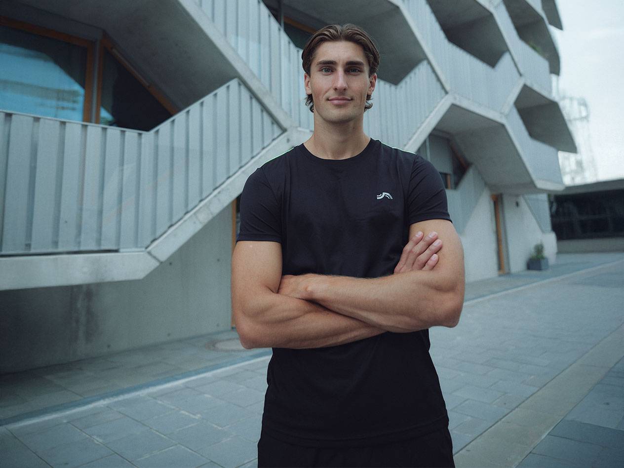 Bărbat în tricou negru cu logo și pantaloni scurți negri, stând afară cu brațele încrucișate.