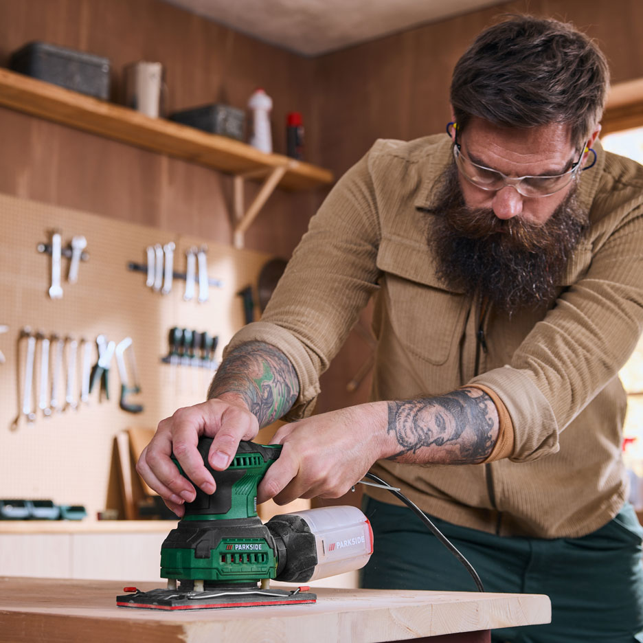 Un bărbat într-un atelier șlefuiește o scândură de lemn cu un șlefuitor electric.