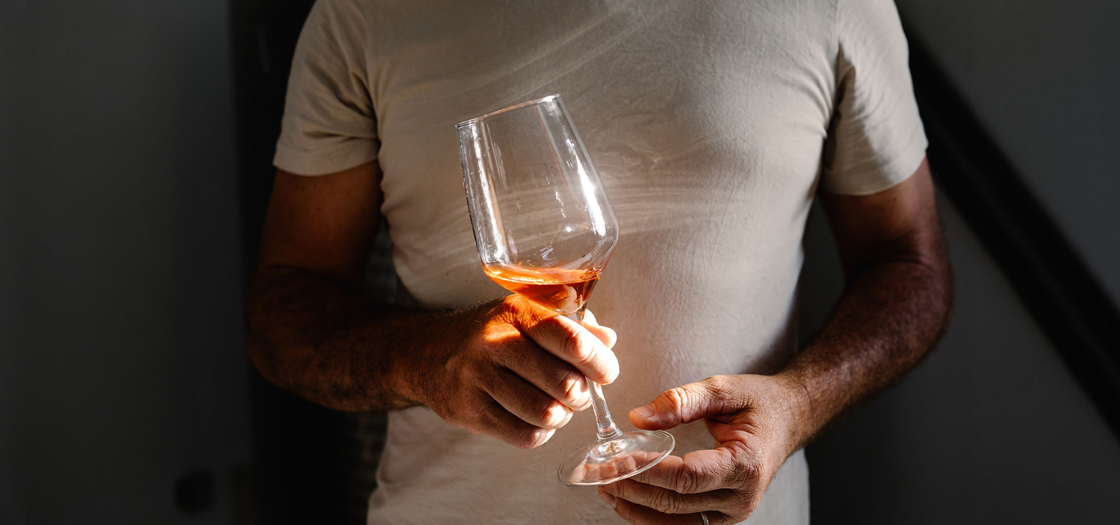 Bărbat ținând un pahar de vin rosé, scăldat în lumina soarelui, pe un fundal întunecat.