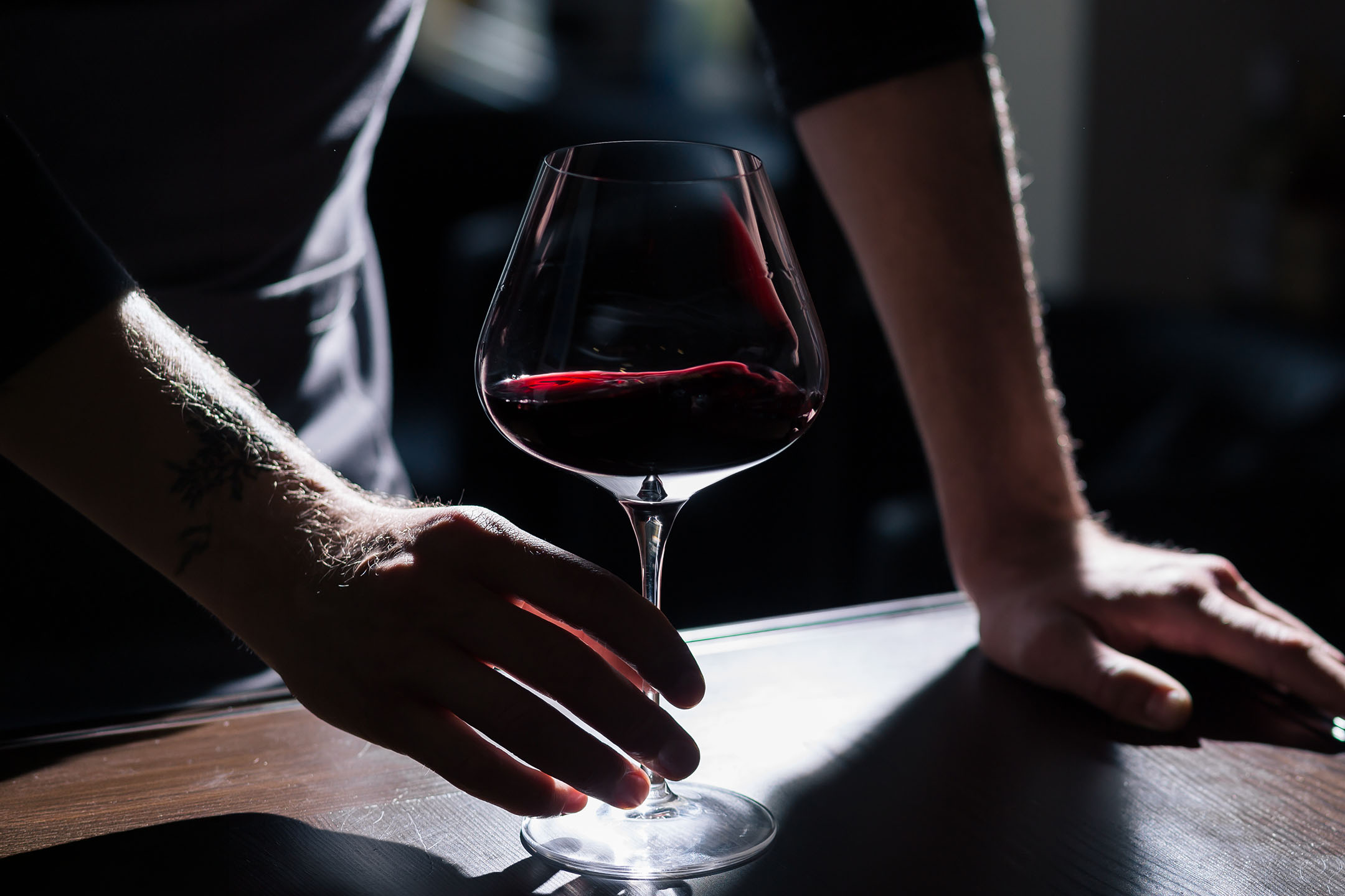 Mâinile tatuate ale unui bărbat țin un pahar de vin roșu pe o masă de lemn.