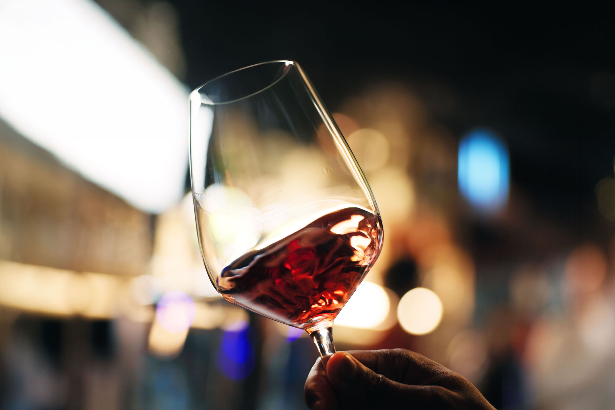 Pahar de vin roșu ținut în mână, cu lumini estompate în fundal.