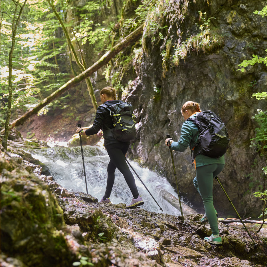 Două femei cu rucsaci și bețe de trekking merg pe lângă o cascadă.