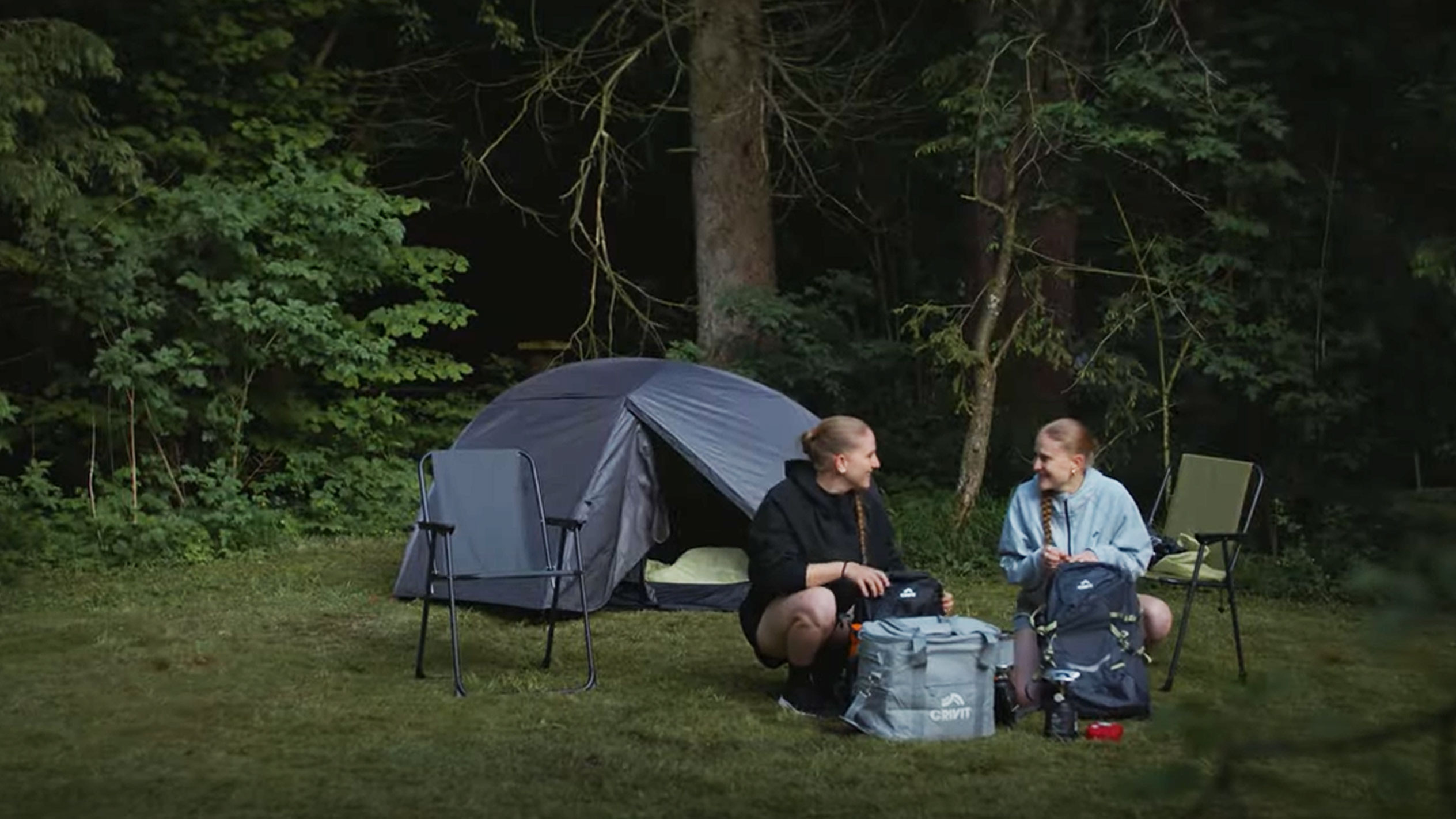 Două femei fac camping cu un cort Crivit și scaune pliante.