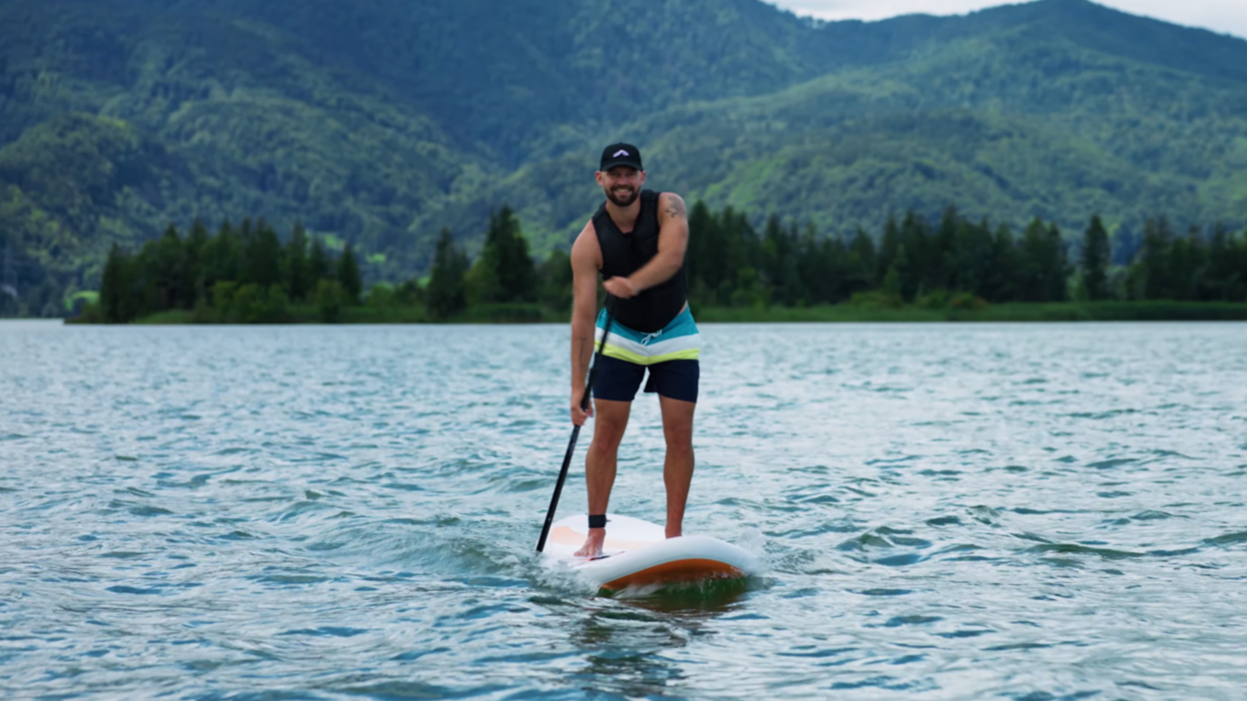 Bărbat pe placă de paddleboard pe un lac cu munți în fundal.