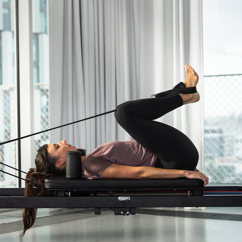 Femeie care face Pilates pe un aparat reformer, cu picioarele ridicate.