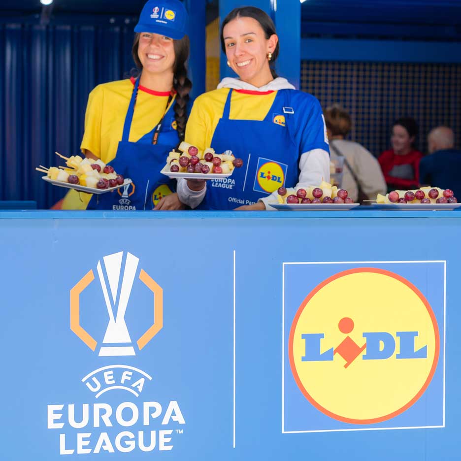 Două femei zâmbitoare în șorțuri Lidl oferă fructe pe bețe la un eveniment UEFA Europa League.