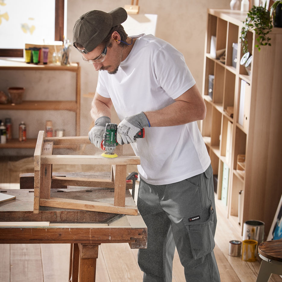 Bărbat șlefuind mobilier din lemn cu o unealtă electrică, purtând pantaloni de lucru și echipament de protecție.