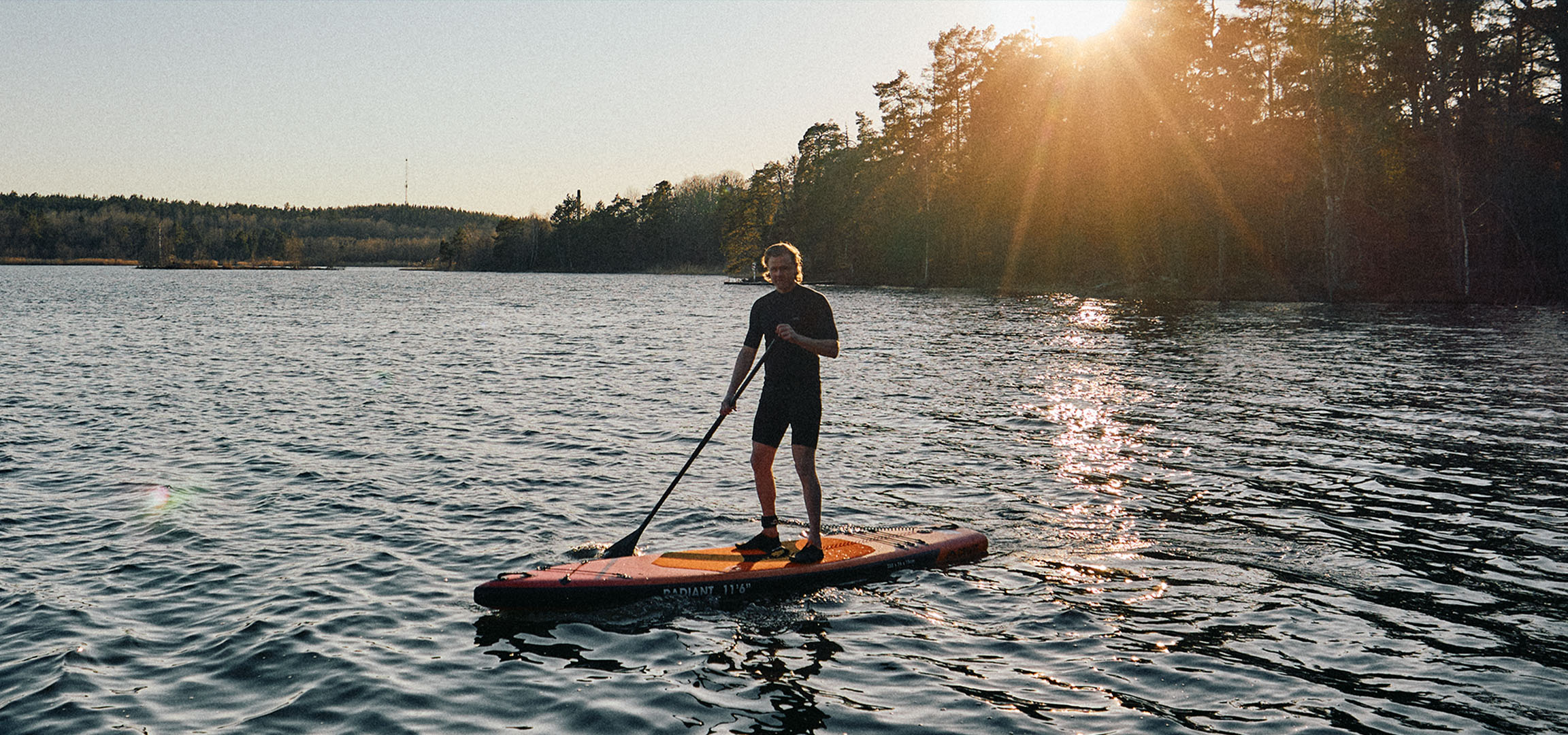 Bărbat pe o placă de stand up paddle pe un lac la apus.