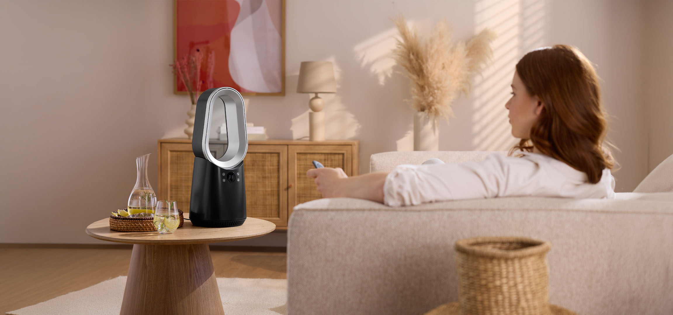 Femeie pe canapea cu telecomandă, ventilator modern și băuturi pe masă.