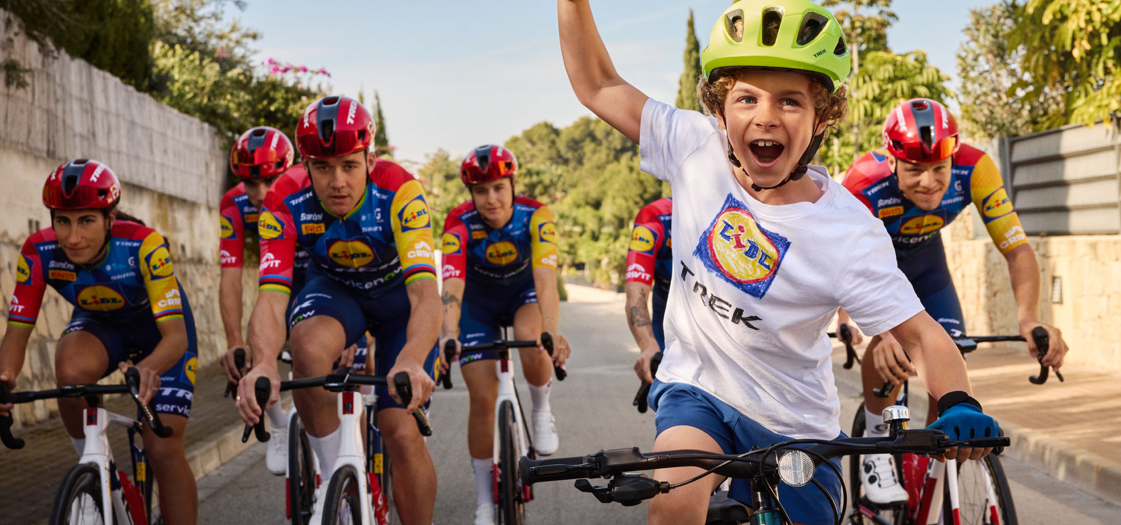 Copil cu cască și tricou cu logo Lidl, înconjurat de cicliști.