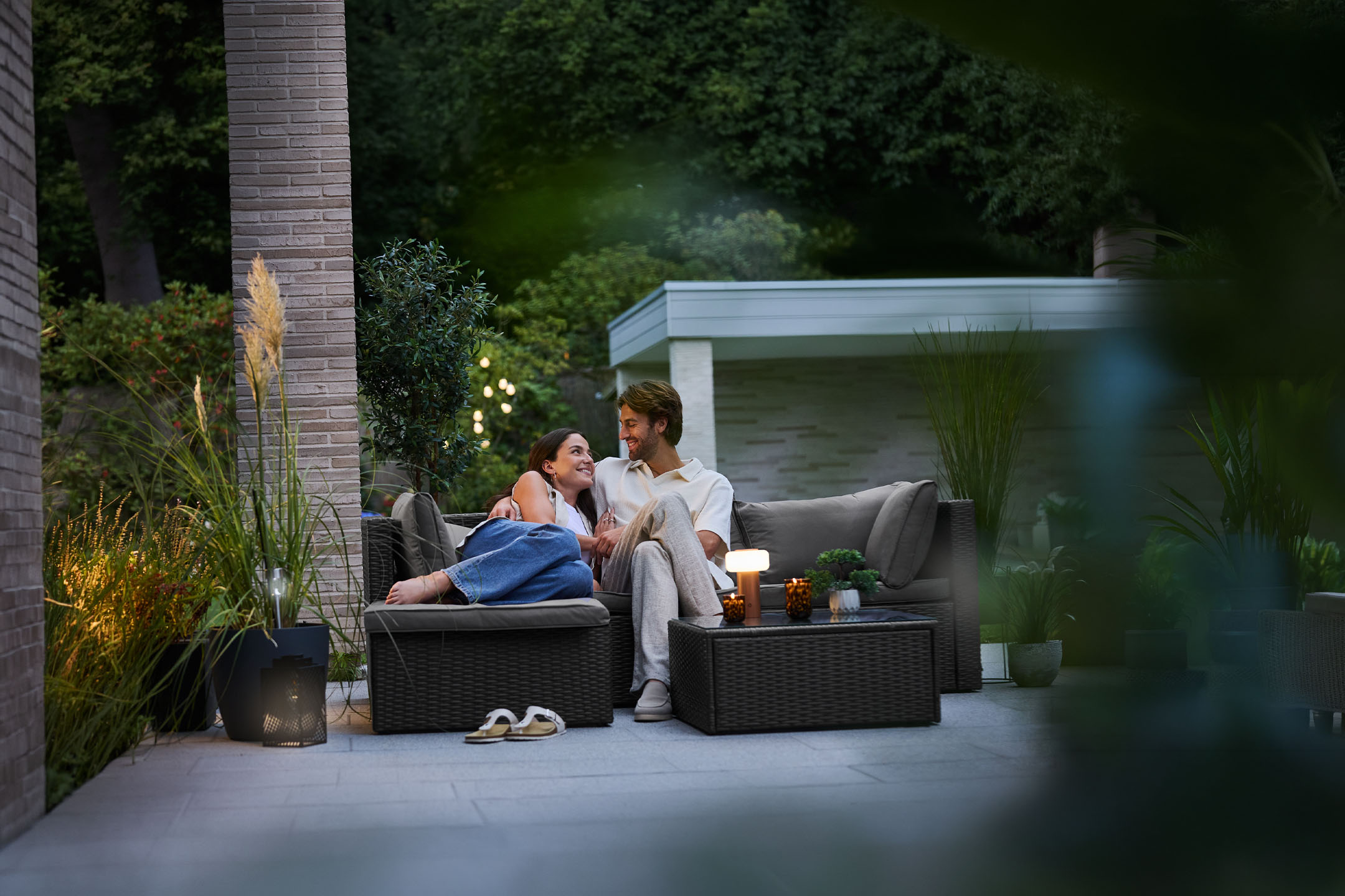 Cuplu relaxându-se pe mobilier de grădină din ratan, iluminat de lămpi de exterior.