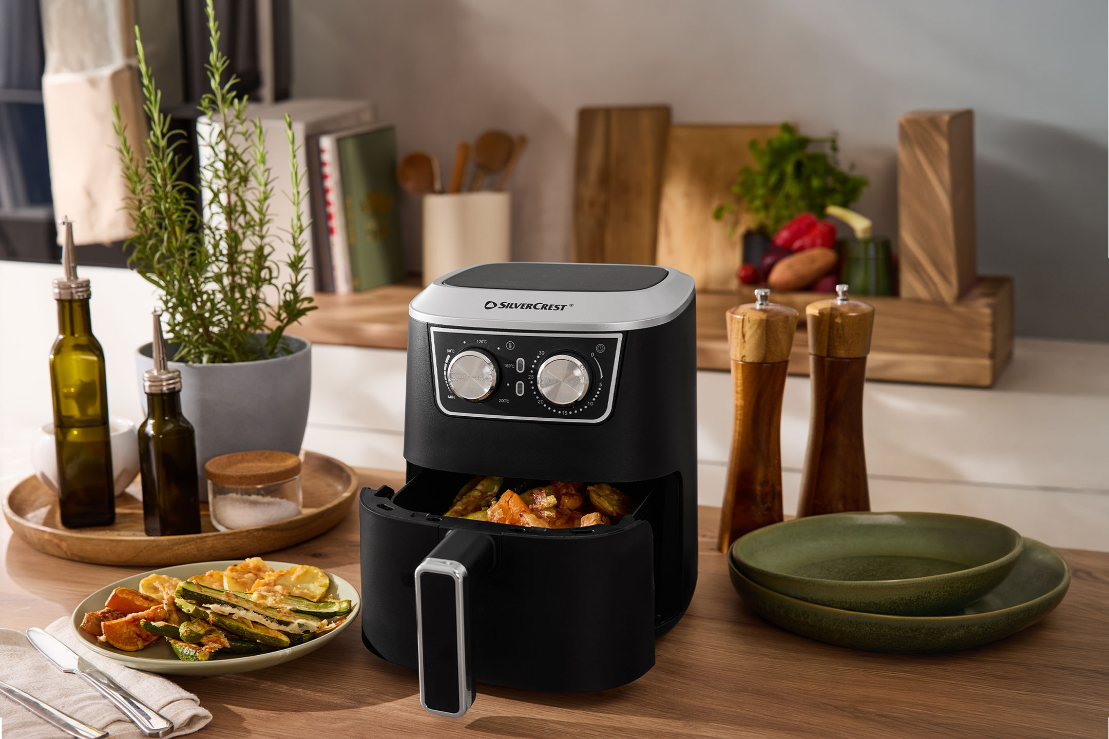 Friteuză cu aer cald neagră cu legume, condimente și vase pe un blat de lemn.