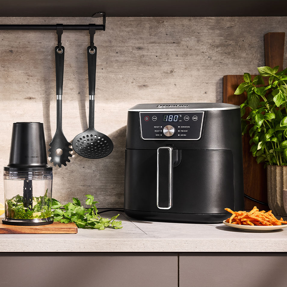 Friteuză cu aer cald SilverCrest și tocător de alimente pe un blat de bucătărie cu ierburi proaspete și cartofi dulci prăjiți.