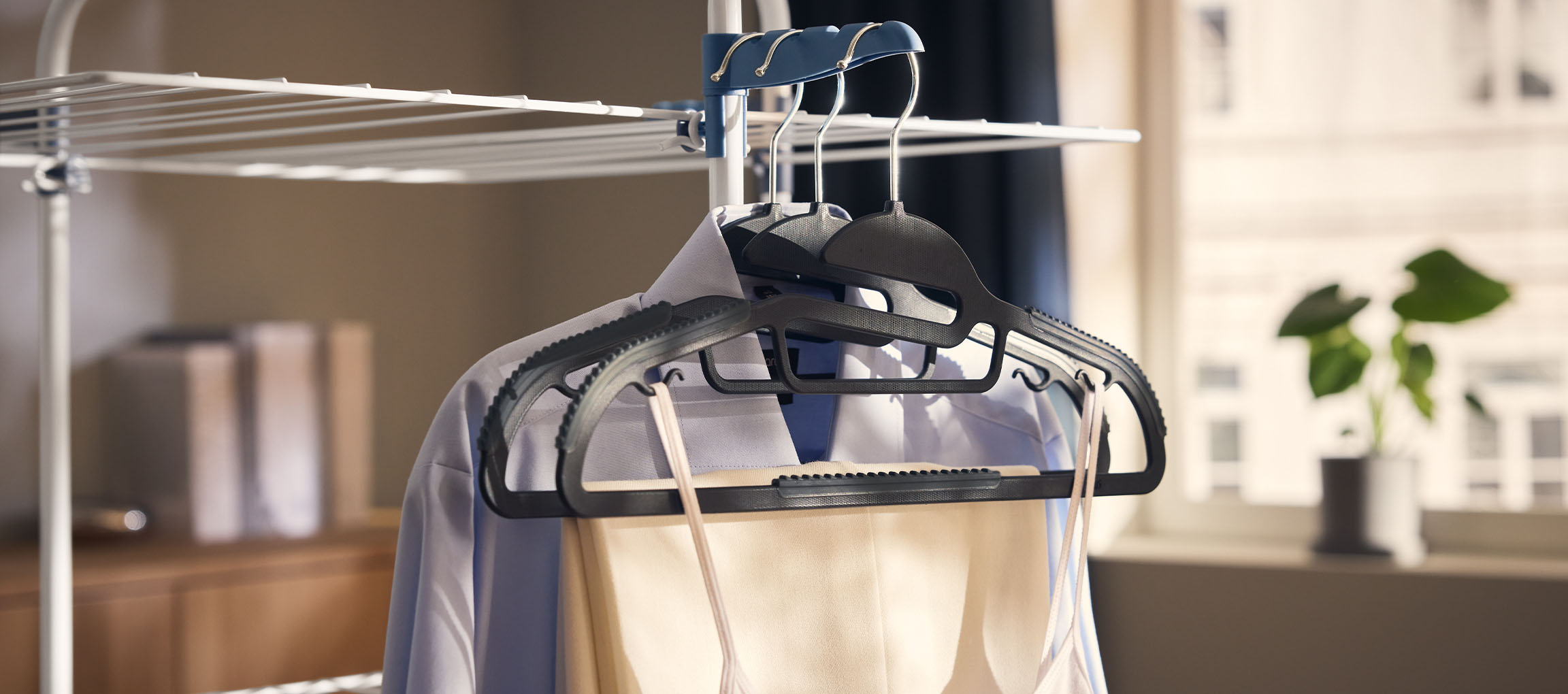 Haine pe umerașe pe un uscător de rufe, cu o plantă în fundal.