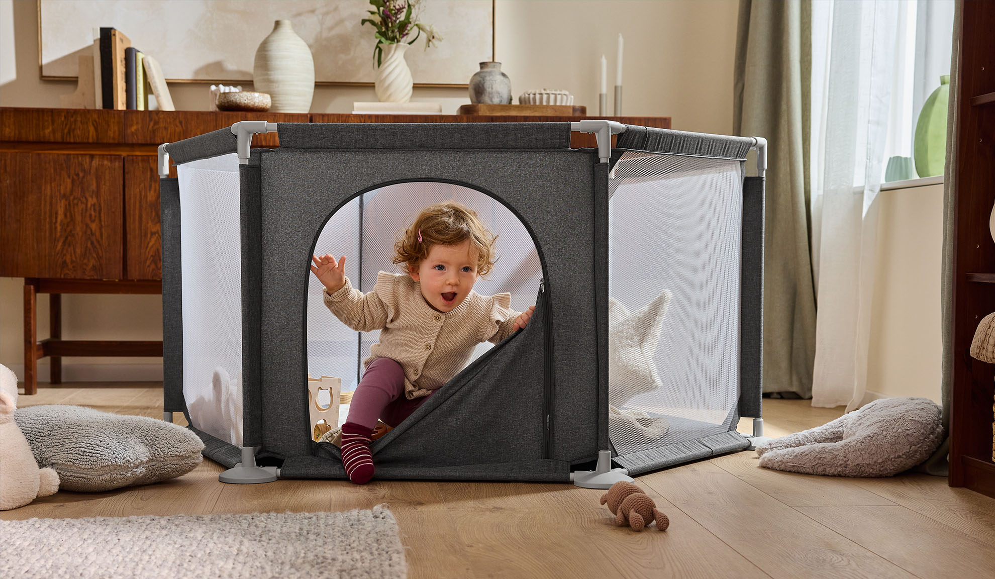 Bebeluș fericit ieșind dintr-un țarc gri într-un living confortabil.