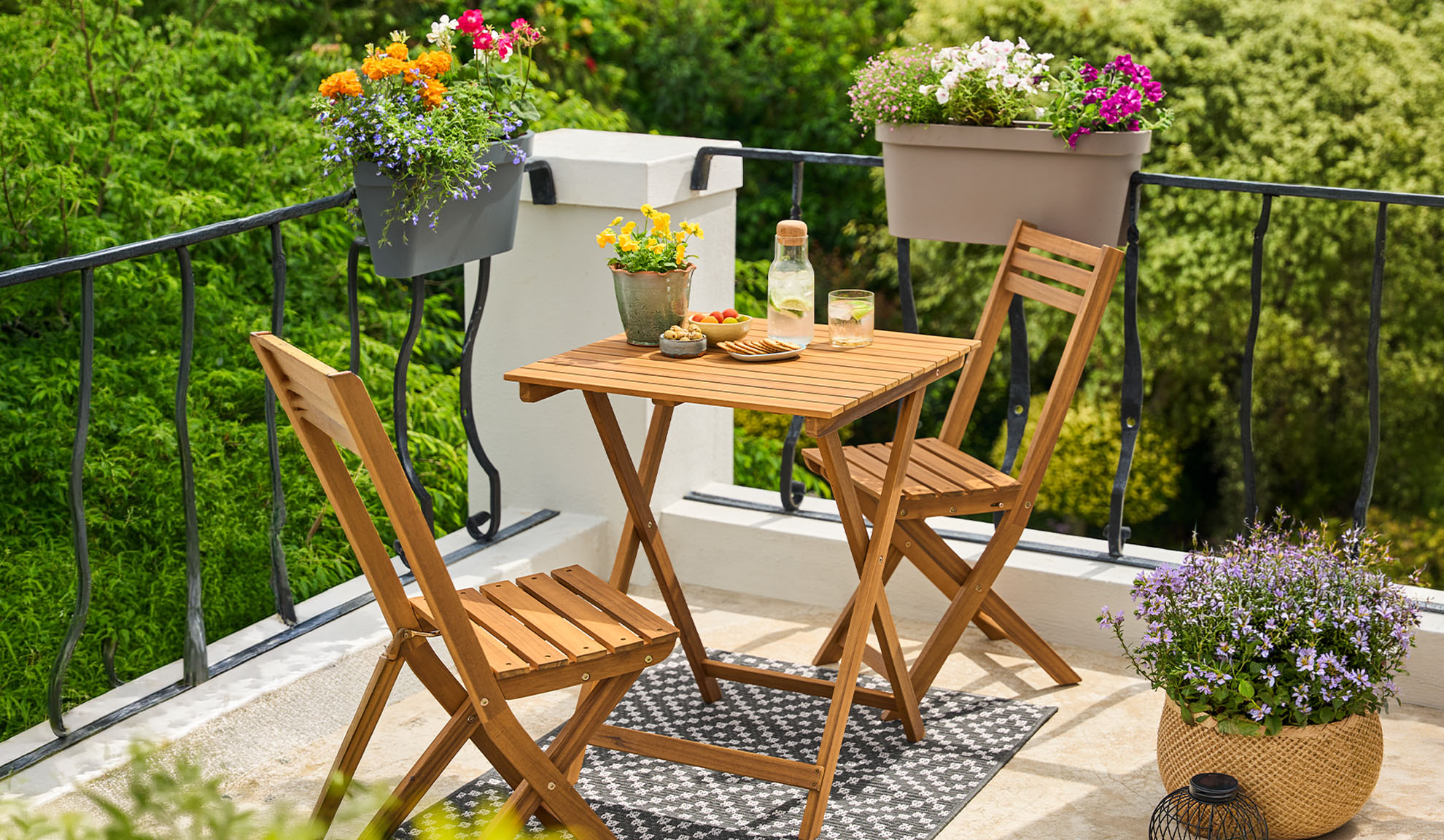 Balcon cu set de bistro din lemn, flori în ghiveci și băuturi pe un covor cu model.