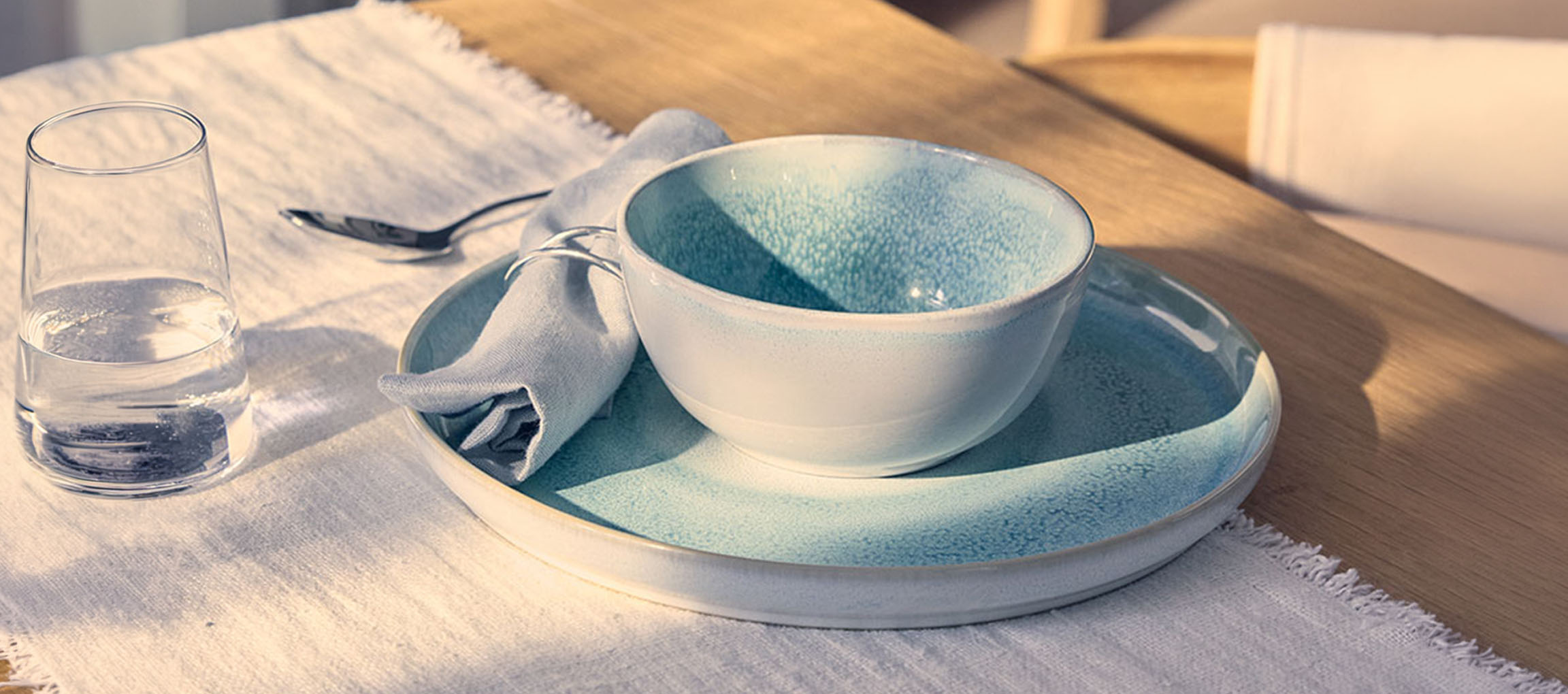 Gedeckter Tisch mit blauer Keramikschale und -teller, Wasserglas und Serviette.