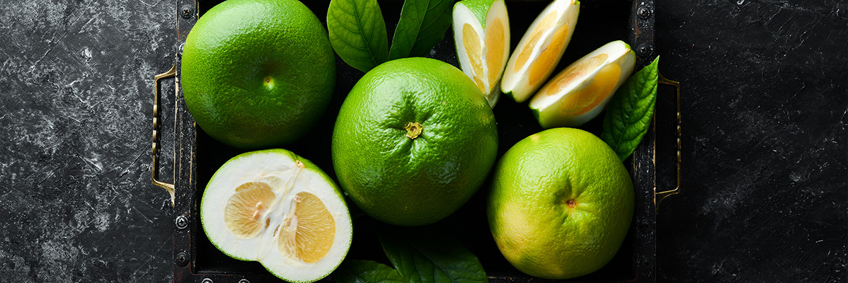 Mai multe grapefruit verzi, unele feliate, într-o tavă de lemn închis la culoare.