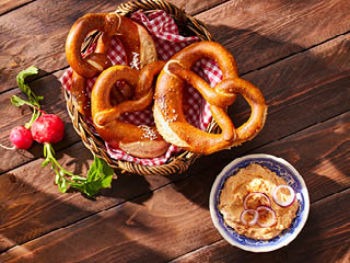 Bretzel proaspeți într-un coș, serviți cu un bol de unt de brânză și ceapă roșie.