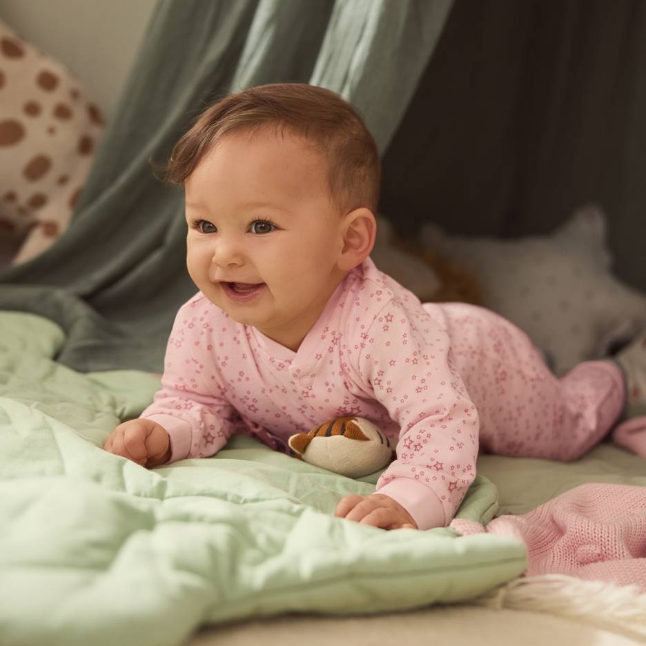Bebeluș în pijama roz cu steluțe, culcat pe o pătură verde cu o jucărie de pluș.
