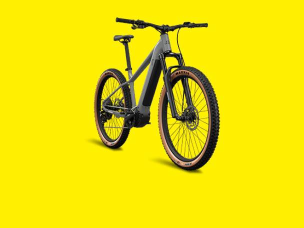 Bicicletă electrică de munte gri cu anvelope maro pe un fundal galben.