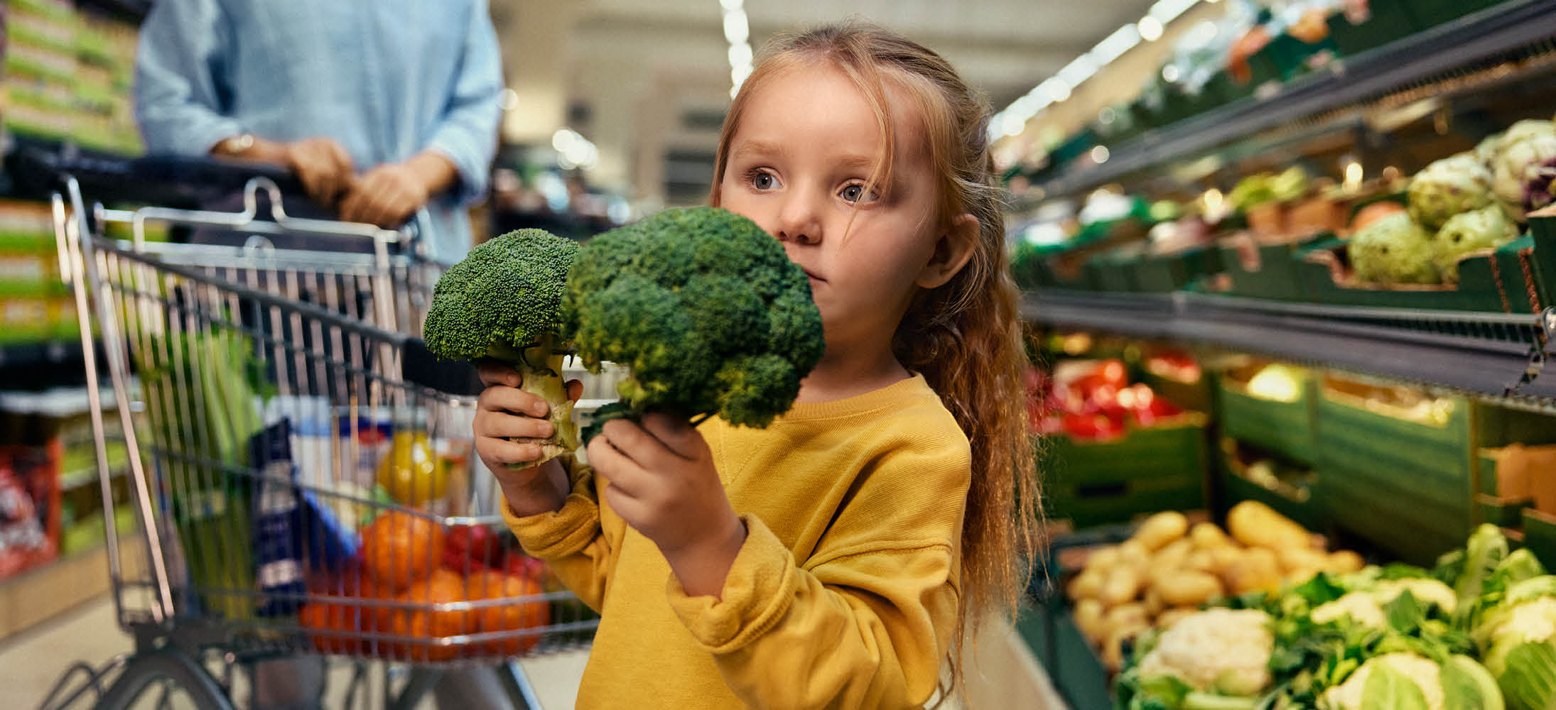 O fetiță ține broccoli în timp ce un adult împinge un cărucior de cumpărături plin cu legume.