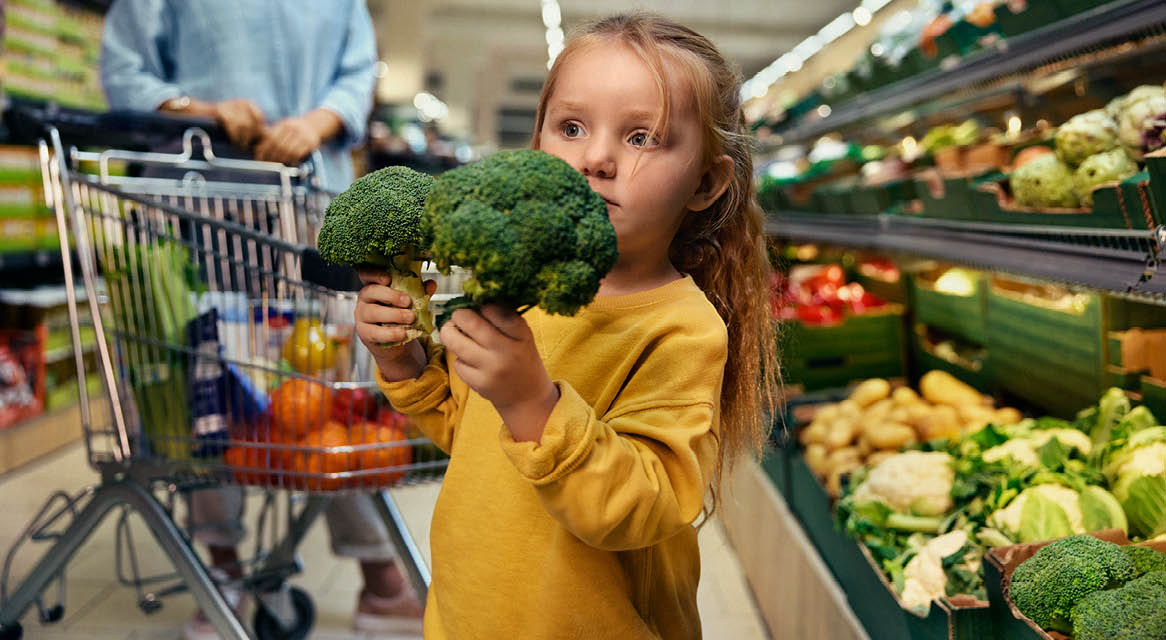 O fetiță ține broccoli în timp ce un adult împinge un cărucior de cumpărături plin cu legume.