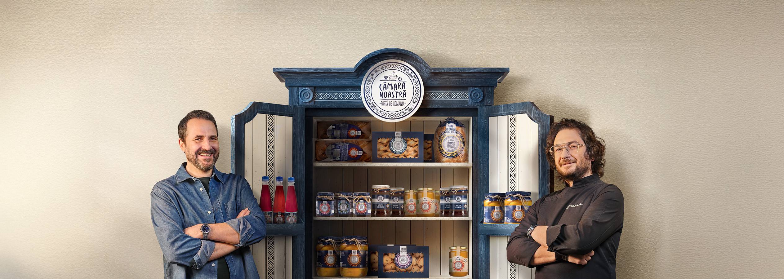Doi bărbați zâmbitori lângă un dulap plin cu produse alimentare „Cămara Noastră”.