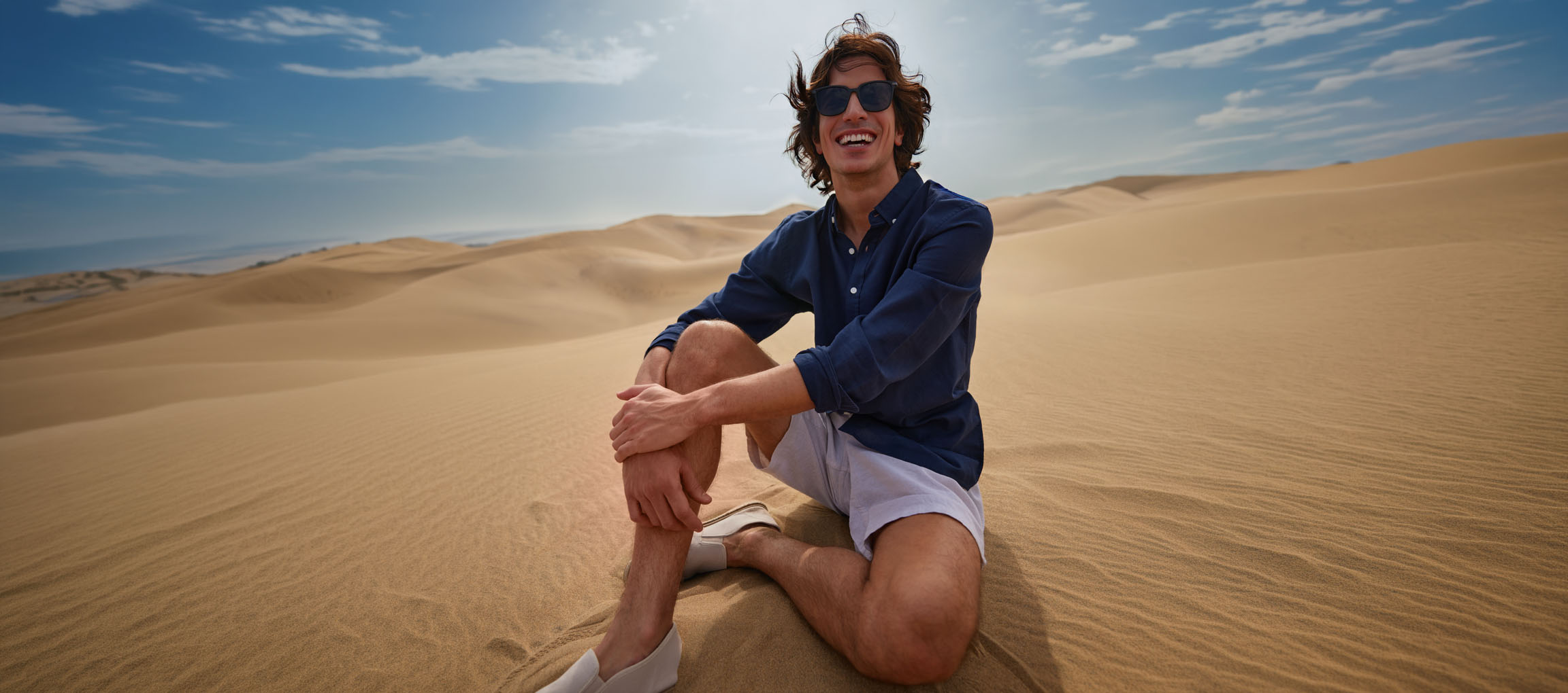Bărbat în cămașă albastru închis și pantaloni scurți stă pe dune de nisip, zâmbind.