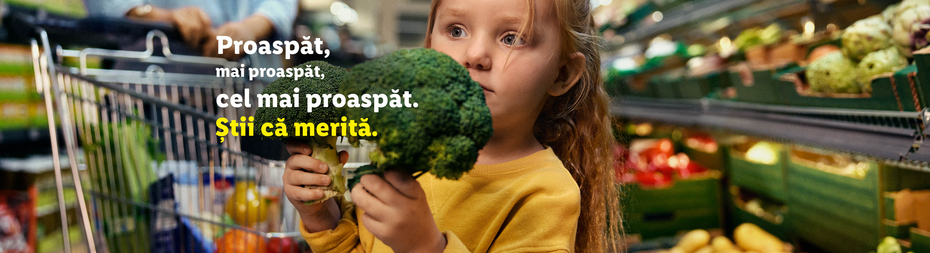 O fetiță ține broccoli în timp ce o femeie împinge un cărucior de cumpărături.