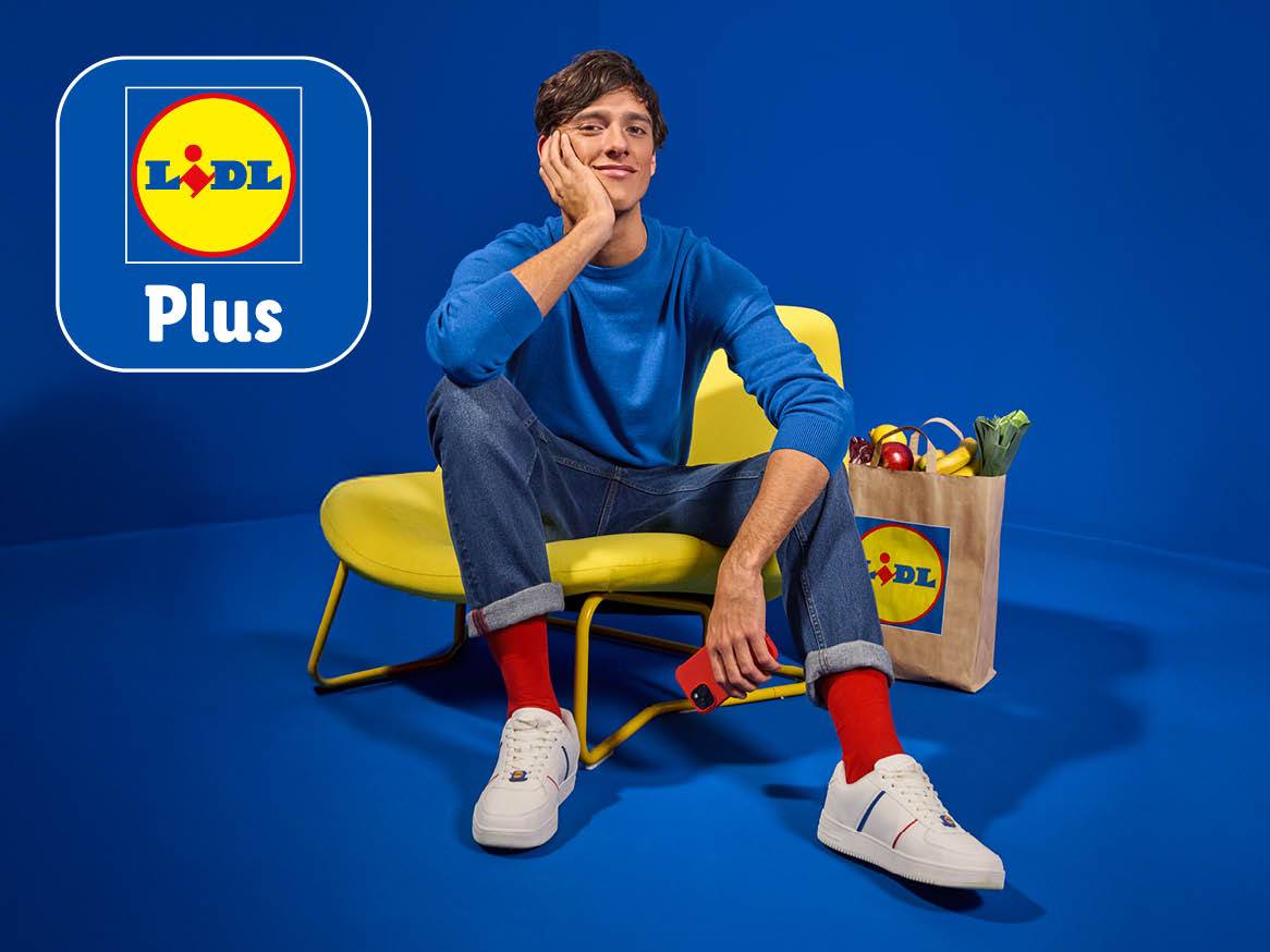 Un bărbat zâmbitor stă pe un scaun galben, cu o pungă de cumpărături Lidl plină cu fructe și legume.