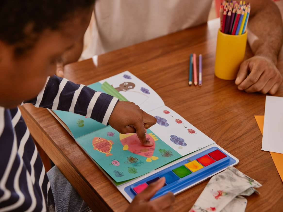 Un copil își folosește degetele pentru a picta o imagine într-o carte de colorat cu ajutorul unor vopsele de acuarelă.
