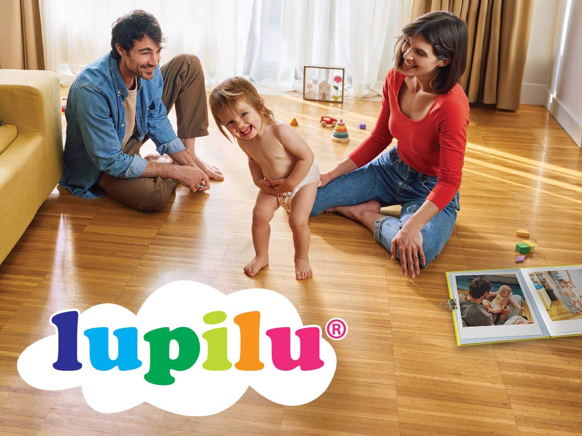 Părinți zâmbitori privesc un copil mic în scutec, cu logo-ul Lupilu.