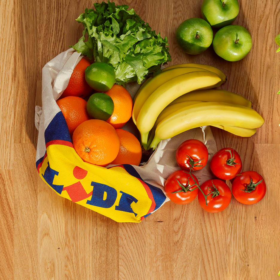 O pungă Lidl plină cu fructe și legume proaspete, inclusiv portocale, banane, salată verde, mere și roșii.