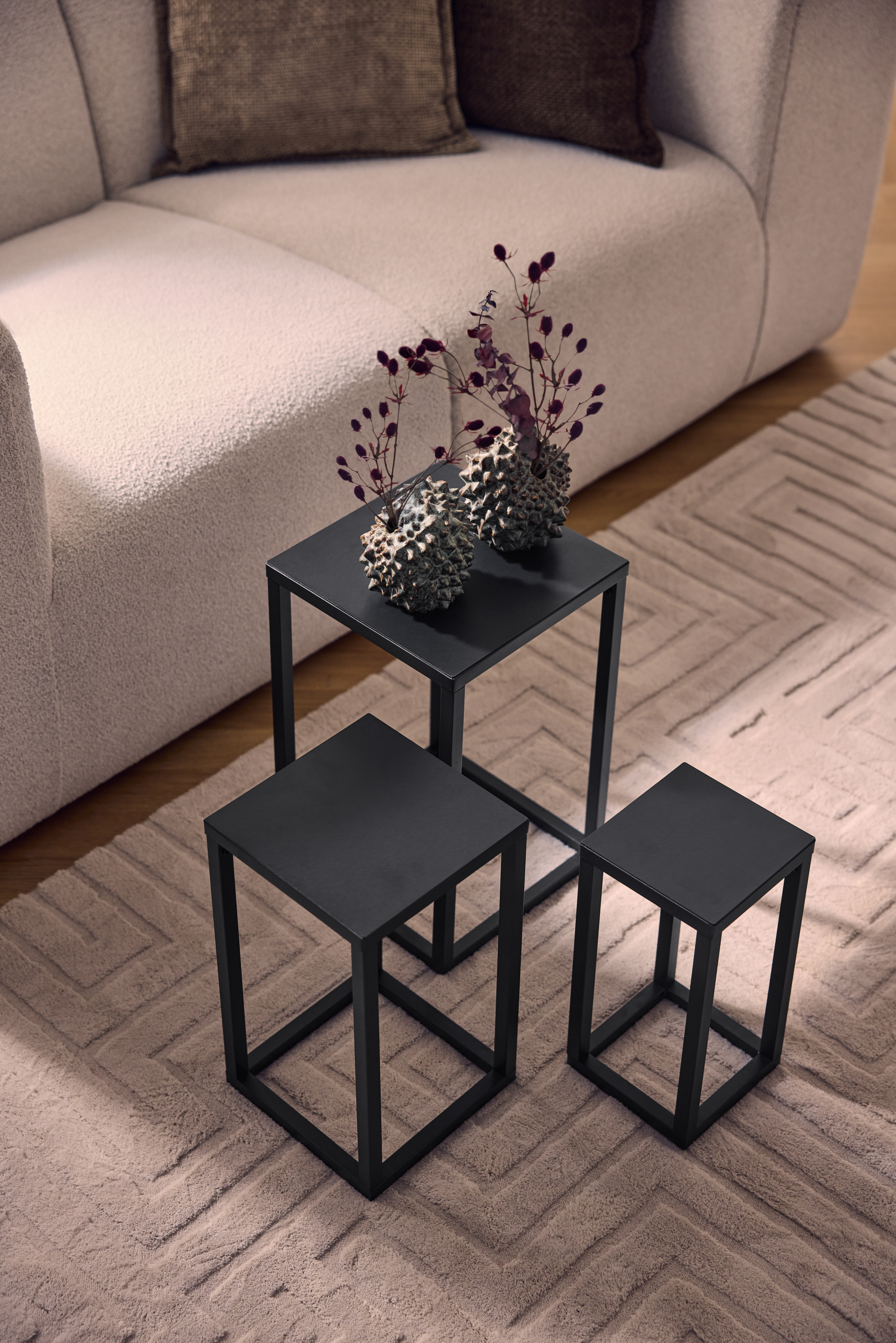 Trei măsuțe de cafea metalice negre pe un covor bej cu model geometric, lângă o canapea deschisă.