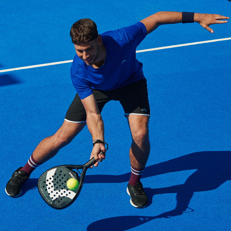 Bărbat în tricou albastru și pantaloni scurți negri jucând padel cu rachetă și minge.