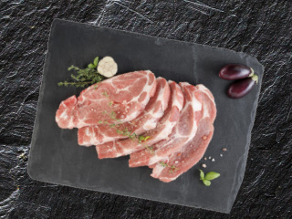 Cotlete de porc crude, feliate, pe o placă de ardezie neagră, cu ierburi și vinete.