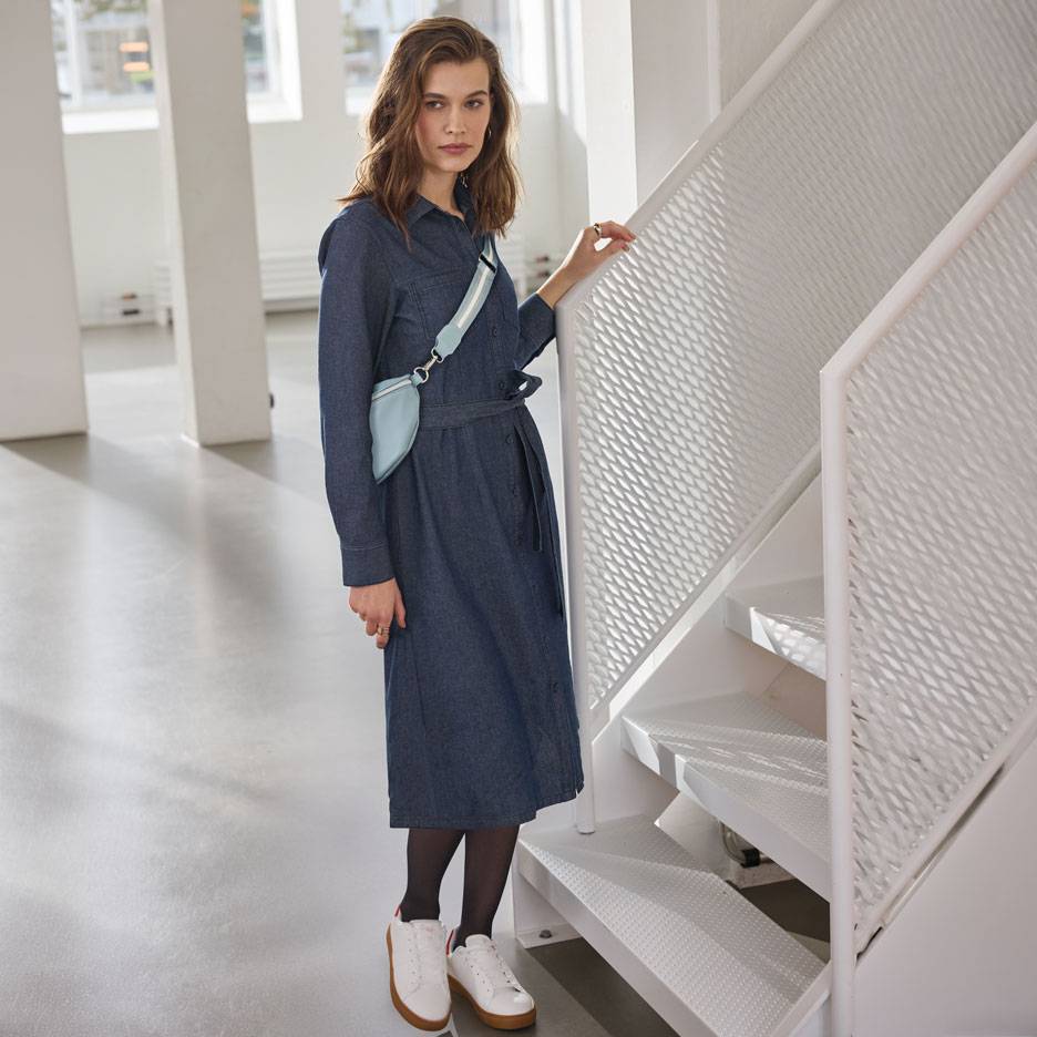 Femeie în rochie cămașă din denim, dresuri negre și adidași albi, cu o borsetă albastru deschis.