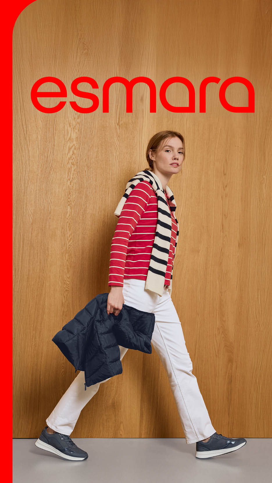 Femeie în tricou roșu cu dungi, pantaloni albi, cu geacă neagră în mână, pe fundal de lemn.