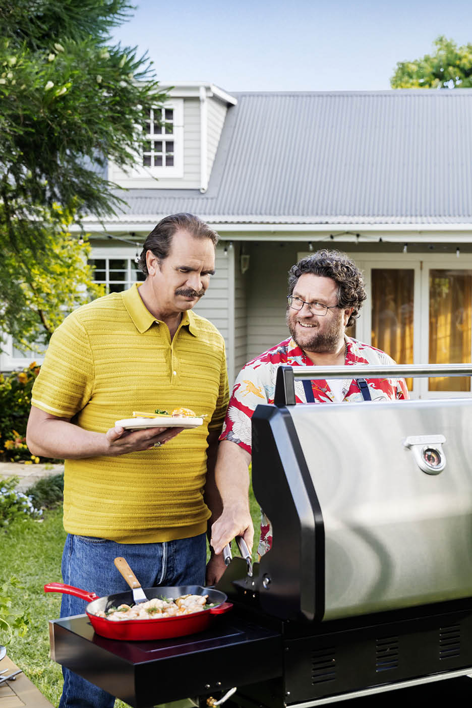 Doi bărbați la grătar, unul ține o farfurie cu mâncare, celălalt gătește într-o tigaie.
