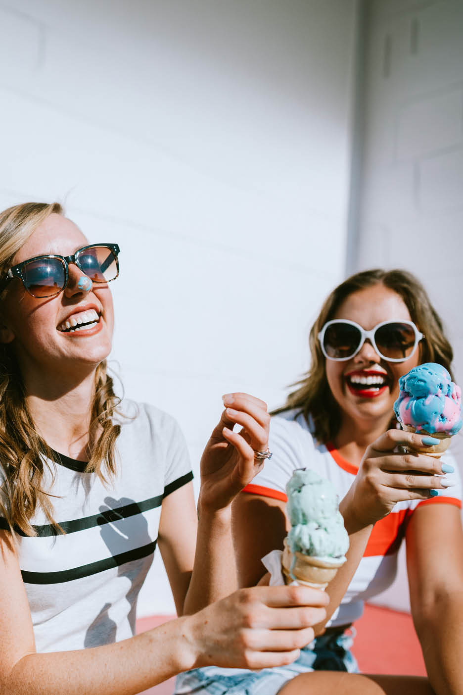 Două femei zâmbitoare cu ochelari de soare, ținând cornete de înghețată colorate.