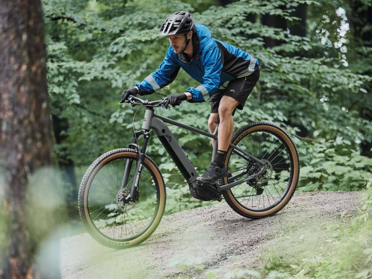 Bărbat pe o bicicletă electrică de munte, purtând o jachetă de ciclism și pantaloni scurți, pe o potecă forestieră.